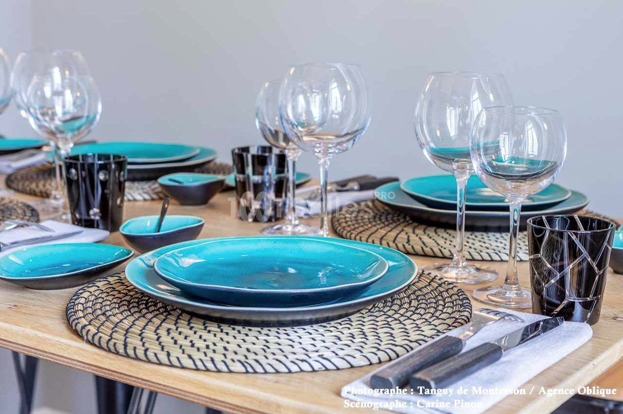 Table dressée avec assiettes turquoise, verres à pied, couverts et accessoires noirs sur un chemin de table tressé.