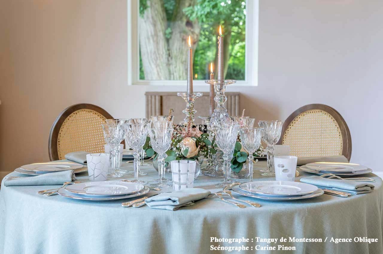 Table élégamment dressée avec nappes bleues, vaisselle raffinée, verres en cristal et bougies allumées au centre.