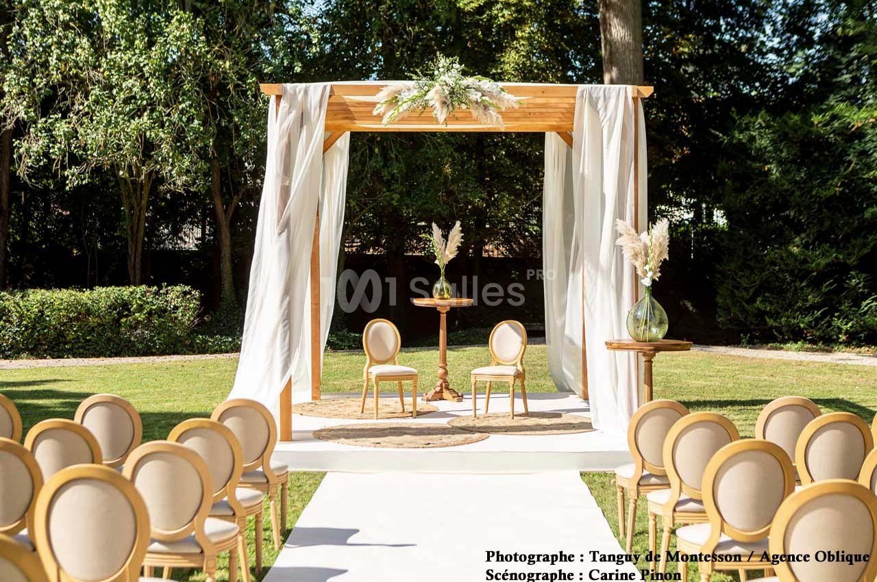 Structure en bois décorée de fleurs blanches, entourée de chaises, installée dans un jardin pour une cérémonie.