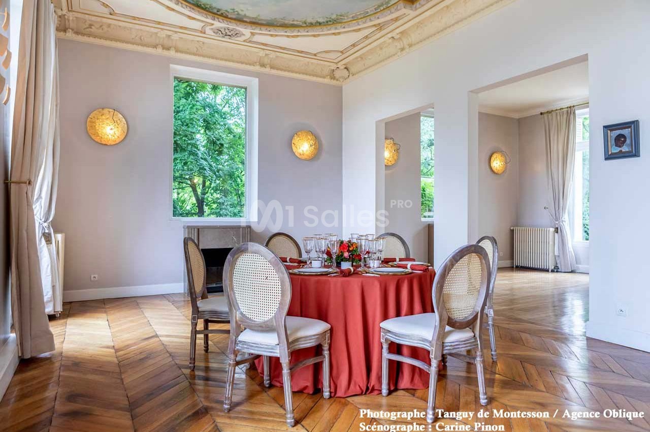 Salle lumineuse avec parquet en bois, table ronde dressée avec nappe rouge et chaises, décorée de moulures au plafond.