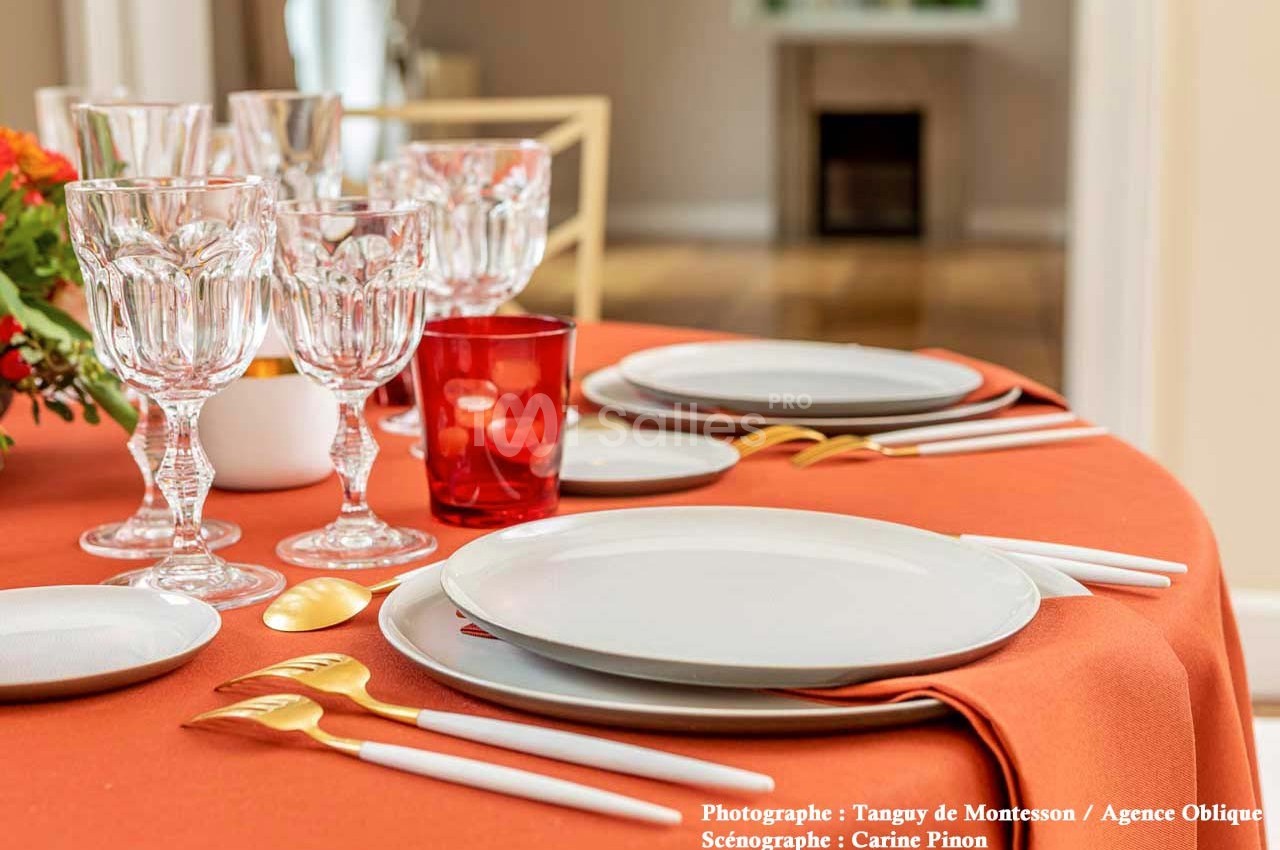 Table dressée avec nappe orange, vaisselle blanche, couverts dorés, verres transparents et un verre rouge en décoration.