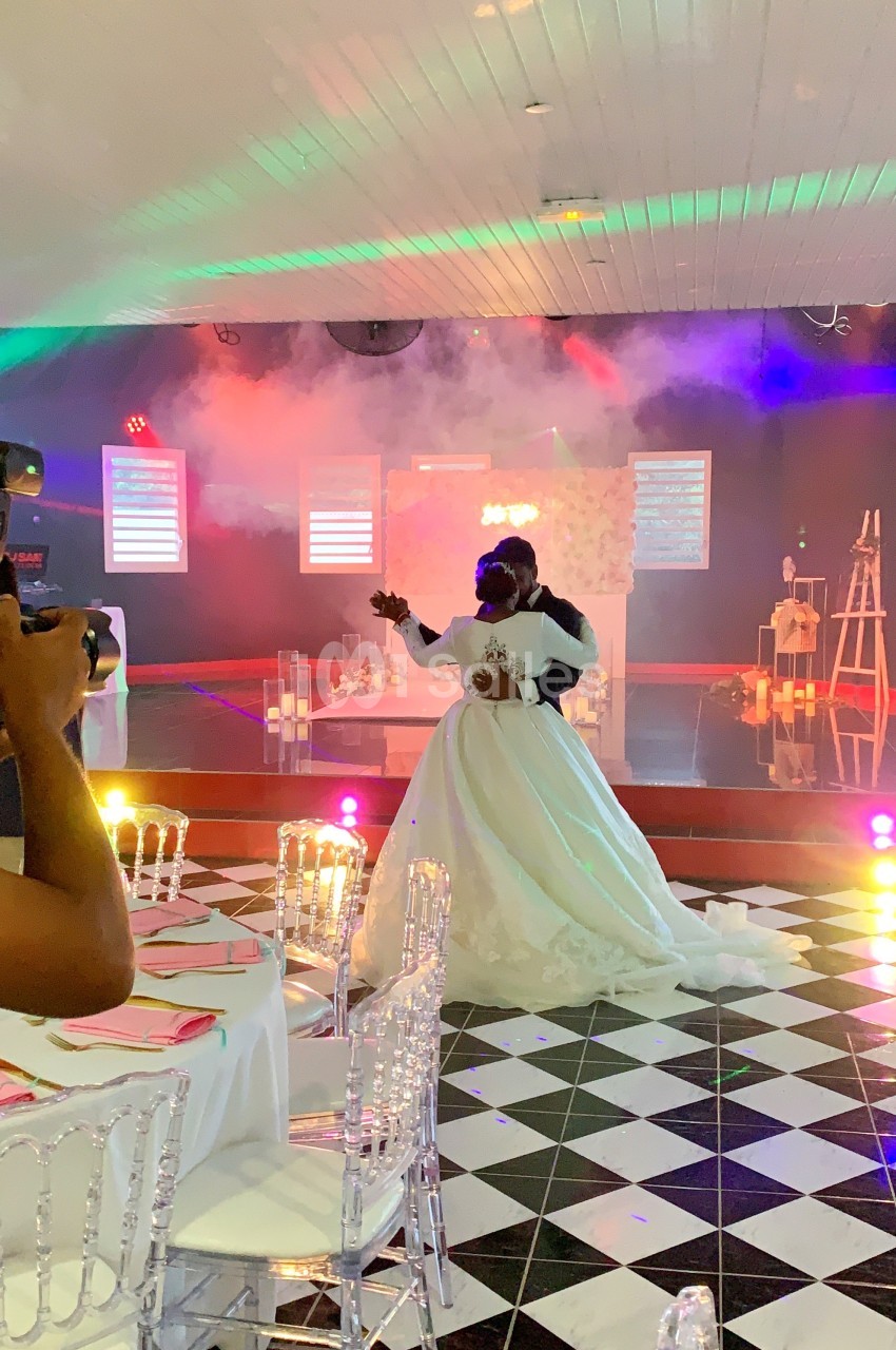 Un couple de mariés danse dans une salle décorée avec des lumières colorées et un sol en damier noir et blanc.