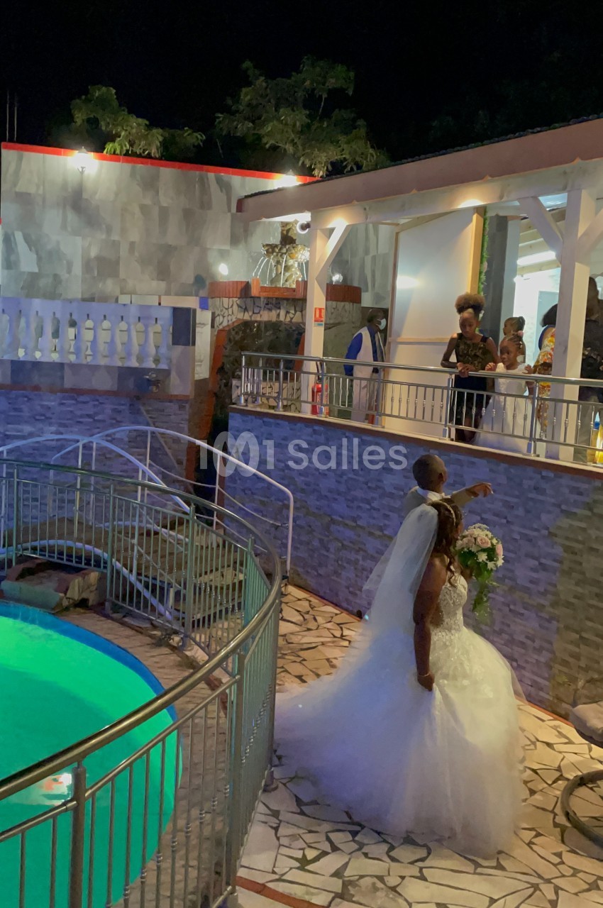 Un couple de mariés pose près d'une piscine éclairée, entouré d'invités sur une terrasse en soirée.