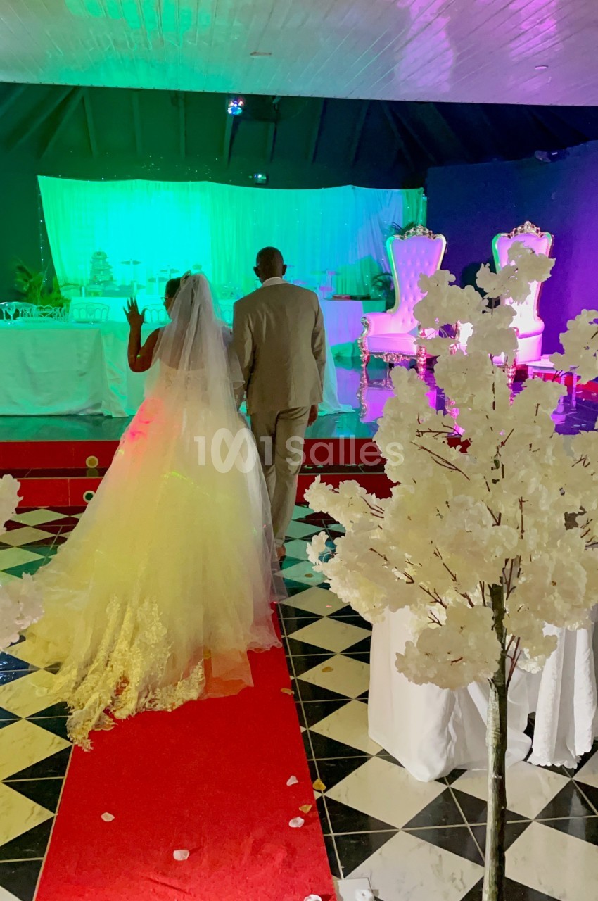 Un couple en tenue de mariage marche sur un tapis rouge dans une salle décorée avec des lumières colorées et des arbres…