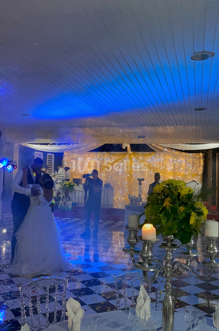 Un couple danse sous des lumières bleues dans une salle décorée avec des guirlandes lumineuses et des fleurs.
