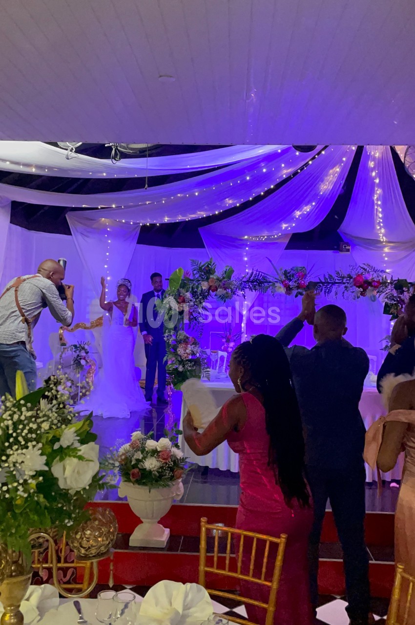 Un couple de mariés pose sur une scène décorée, entouré d'invités prenant des photos dans une salle ornée de fleurs et de…