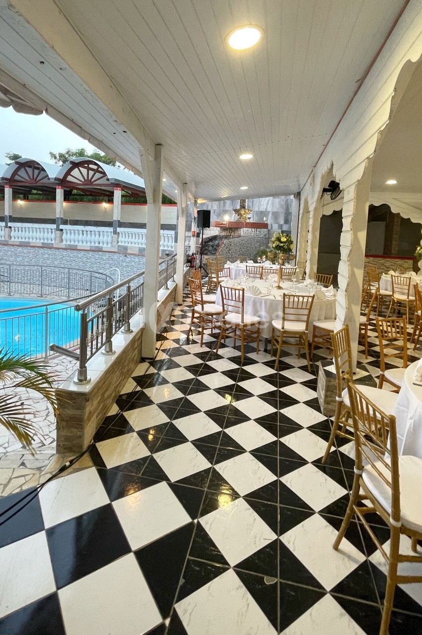 Terrasse couverte avec sol en damier, tables dressées pour un événement et vue sur une piscine extérieure.