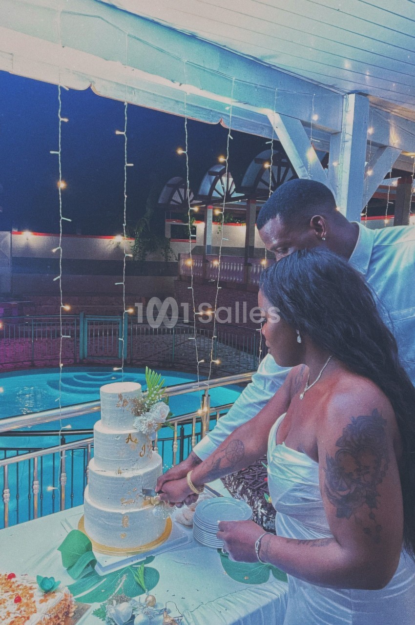 Un couple coupe un gâteau de mariage blanc à plusieurs étages, sous des lumières suspendues près d'une piscine.