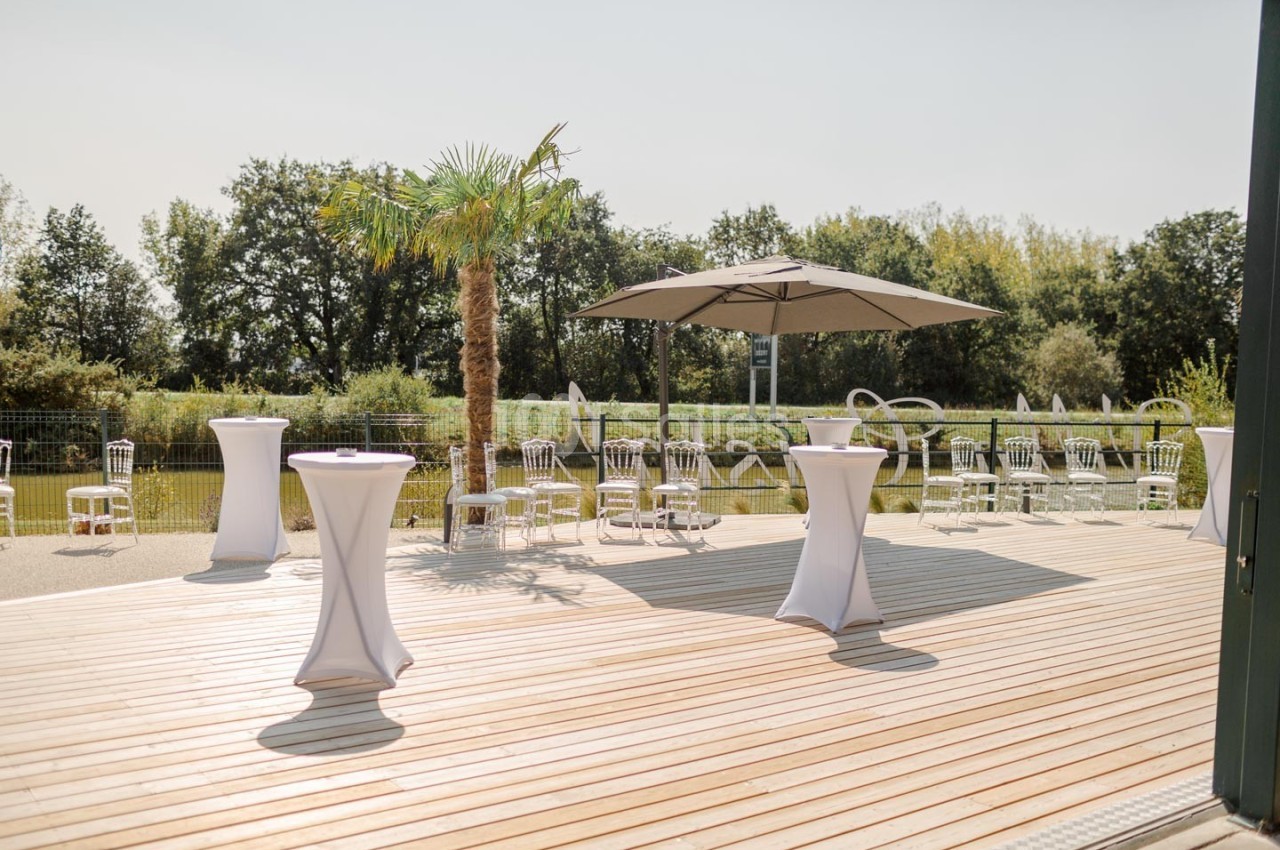 Terrasse ensoleillée avec tables hautes drapées de blanc, chaises et parasol, entourée de verdure.