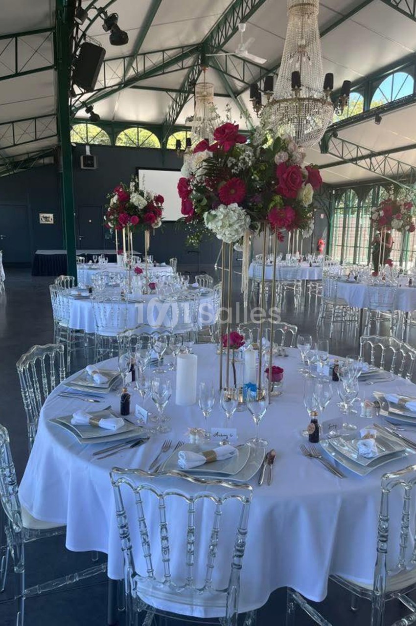 Salle de réception élégante avec tables rondes dressées, décorations florales rouges et blanches, et chandeliers suspendus.