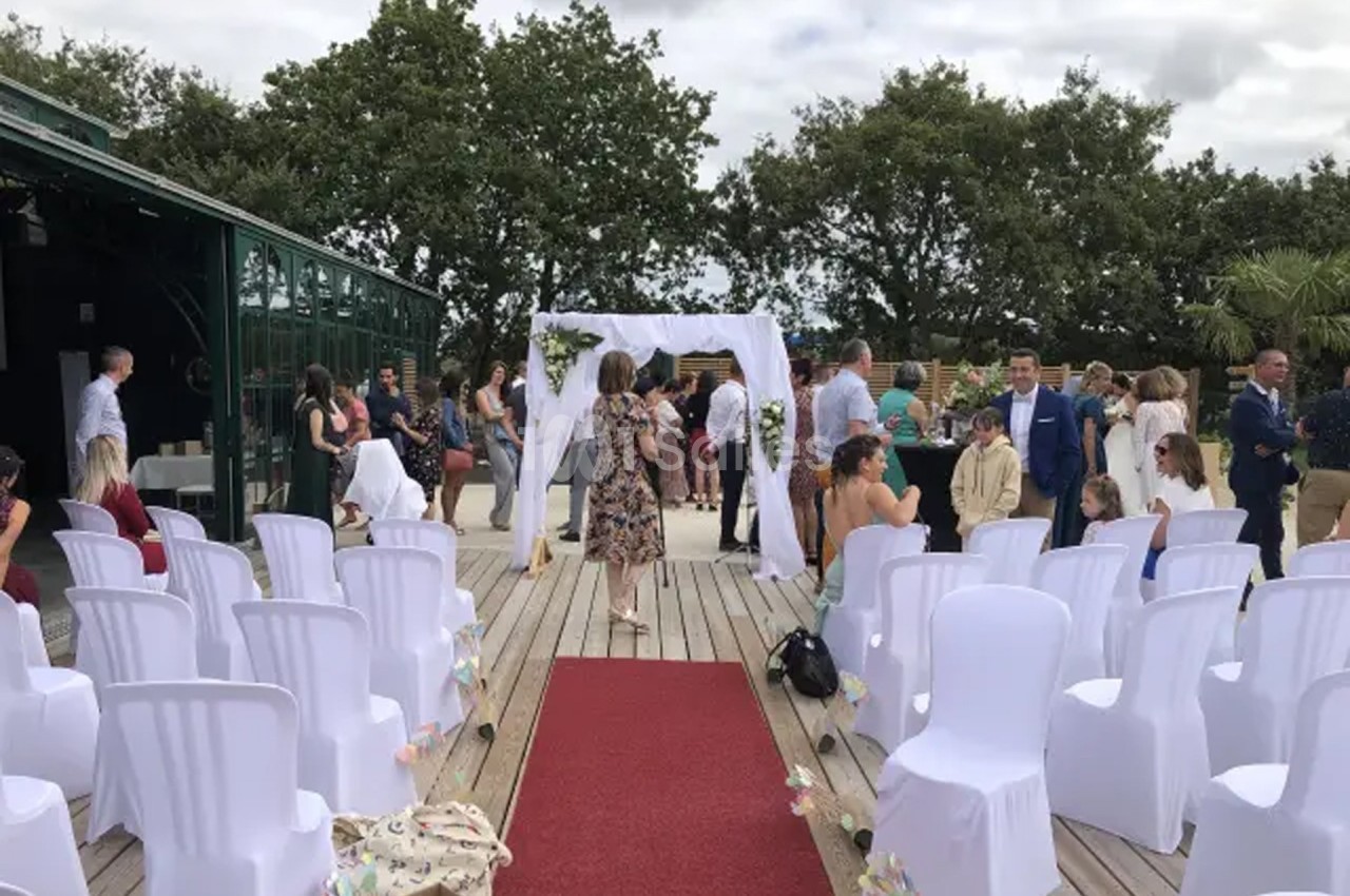 Chaises blanches alignées devant une arche décorée pour une cérémonie en extérieur, avec des invités en arrière-plan.