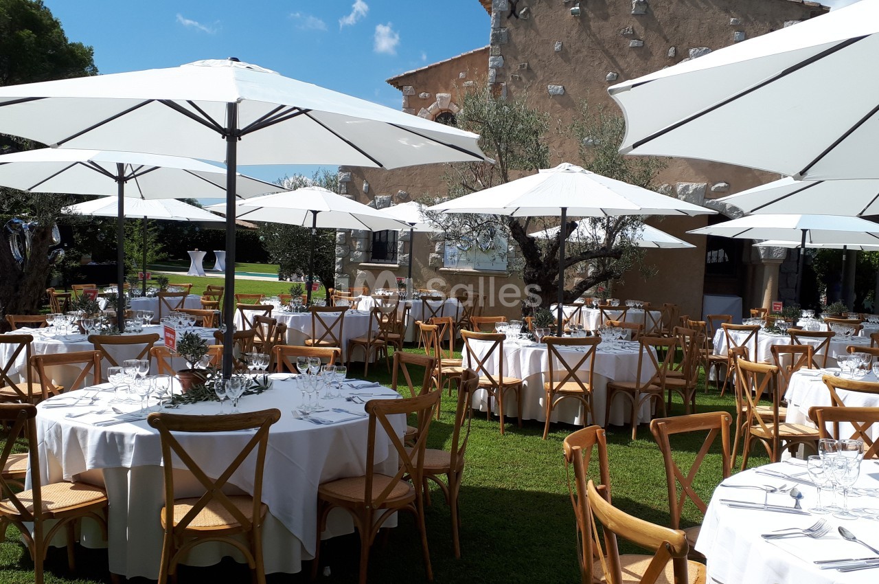 Tables rondes dressées avec nappes blanches et chaises en bois sous des parasols blancs dans un jardin ensoleillé.