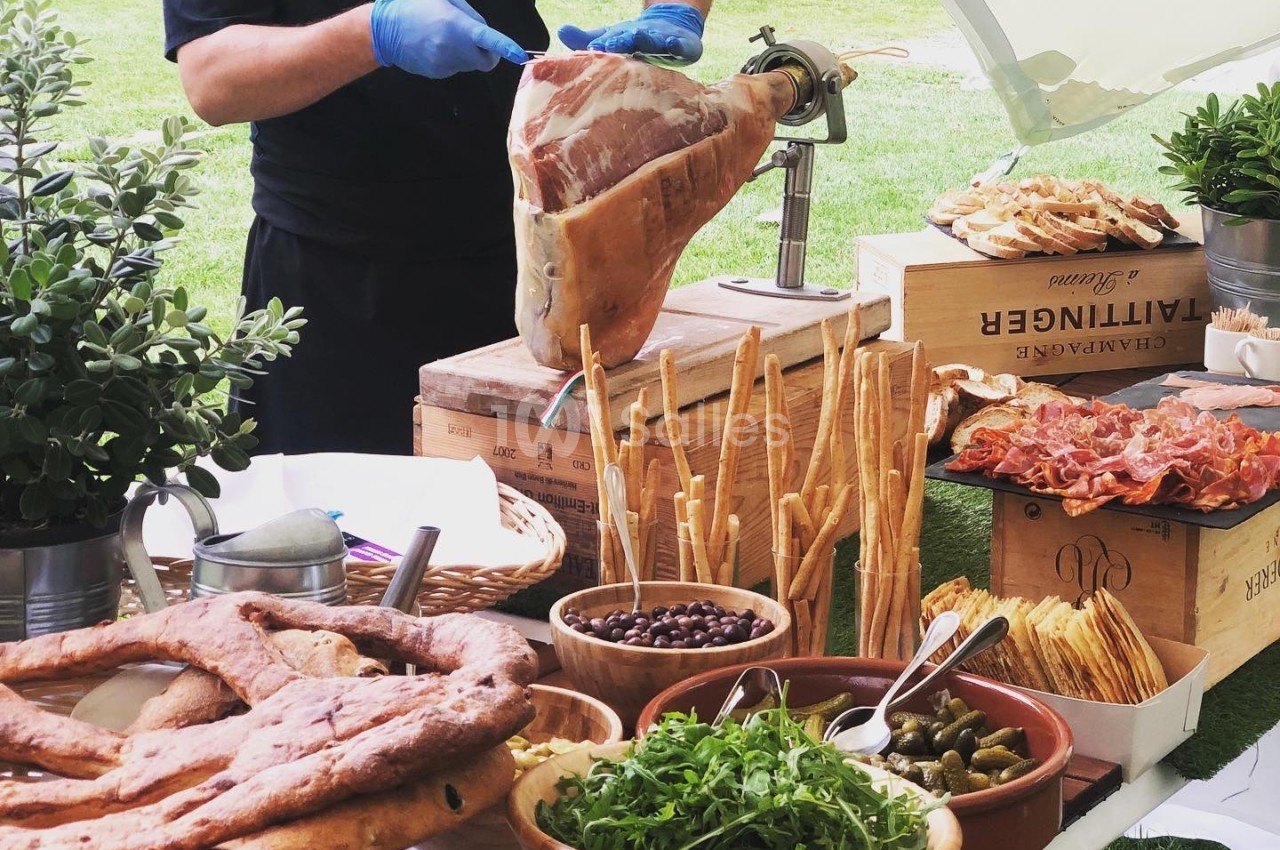 Un chef découpe un jambon cru sur un buffet garni de pains, légumes, olives et charcuteries en extérieur.