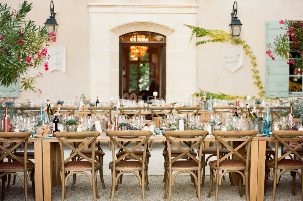 Tables en bois dressées pour un repas en extérieur devant une façade claire avec volets verts et plantes fleuries.
