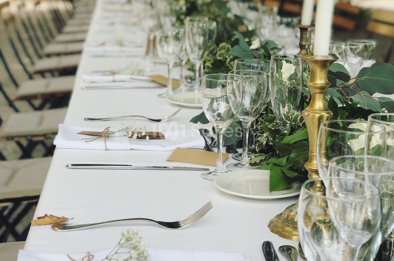 Table dressée pour un repas en extérieur avec nappes blanches, couverts, verres, bougies et décoration florale.