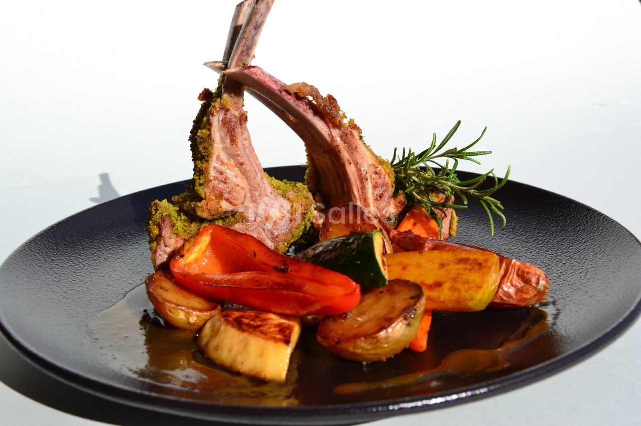 Carré d'agneau en croûte d'herbes servi avec légumes rôtis sur une assiette noire.