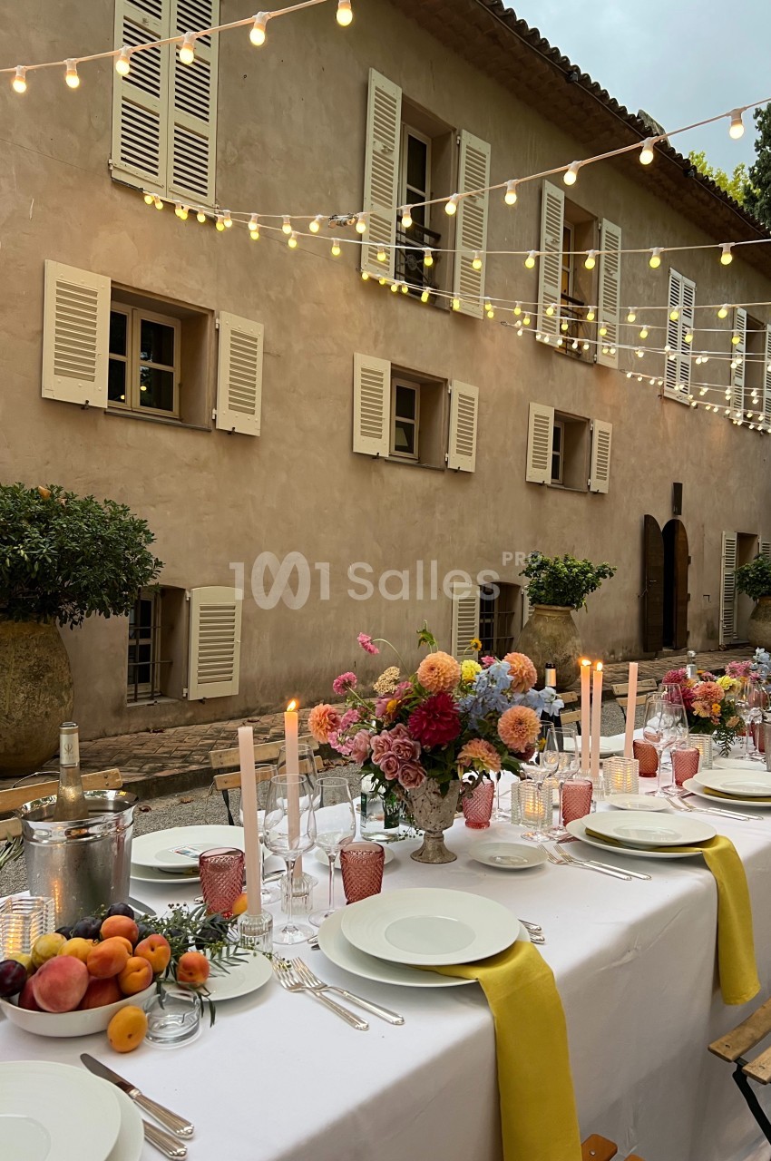 Table dressée pour un dîner en extérieur, décorée de fleurs, bougies et guirlandes lumineuses devant une maison en pierre.