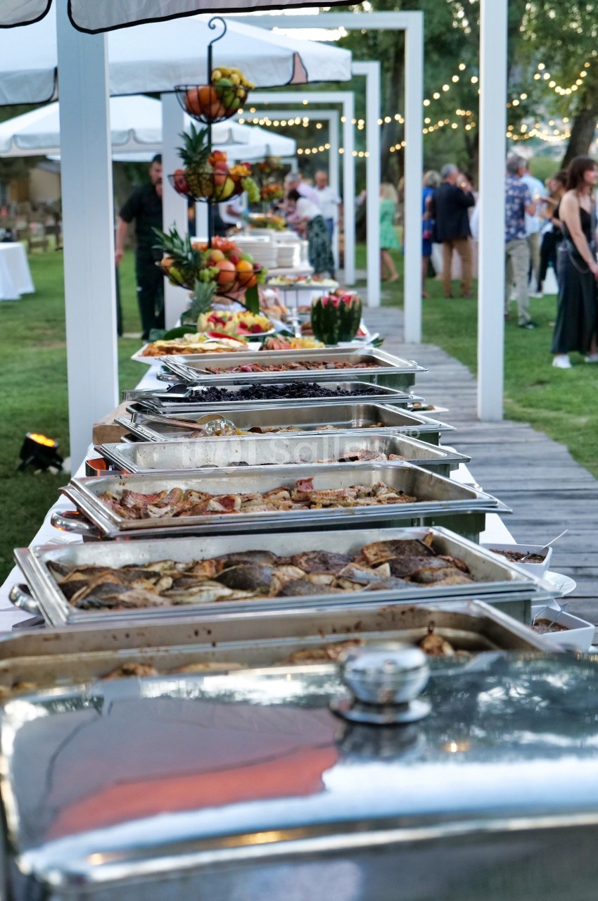Buffet en plein air avec divers plats chauds et froids, installé sur une table dans un jardin décoré de guirlandes…