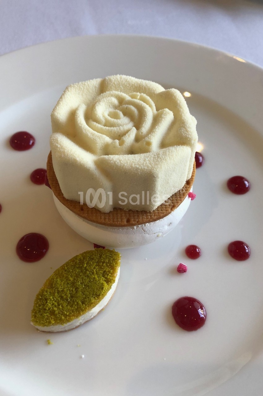 Dessert en forme de rose blanche sur une base de meringue, accompagné de gouttes de coulis rouge et d'une touche verte.