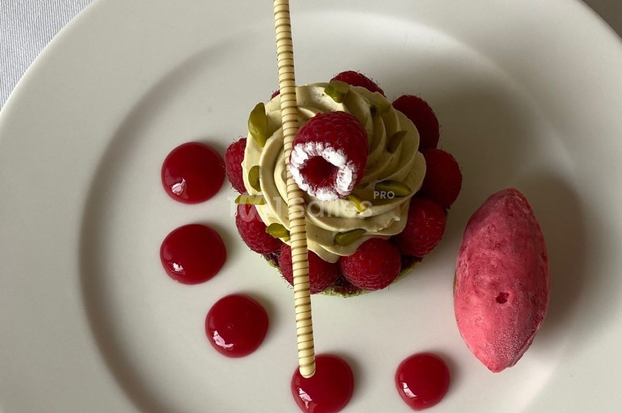 Dessert composé de framboises, crème pistache, décor chocolat, coulis rouge et sorbet framboise sur assiette blanche.