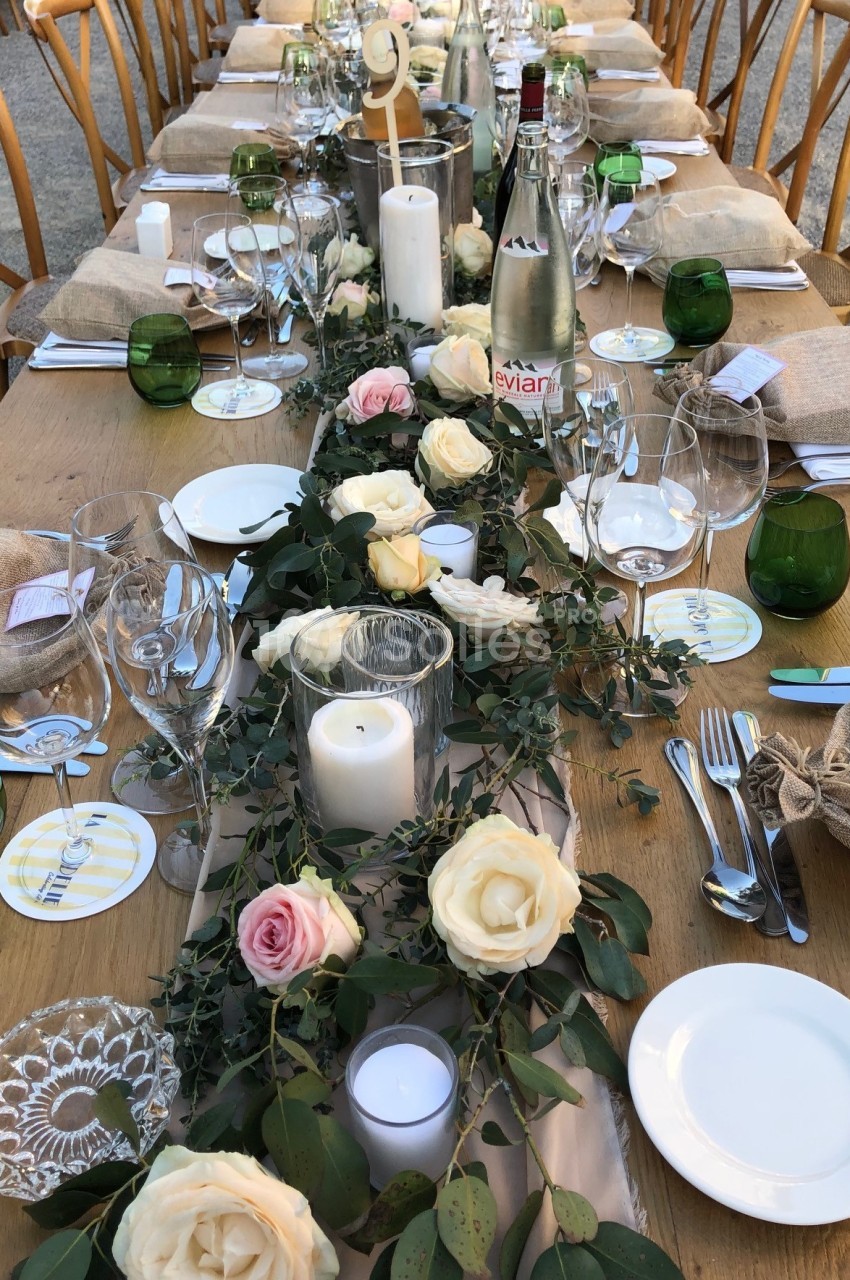 Table en bois décorée pour un repas, ornée de roses, feuillage, bougies blanches et vaisselle disposée avec soin.