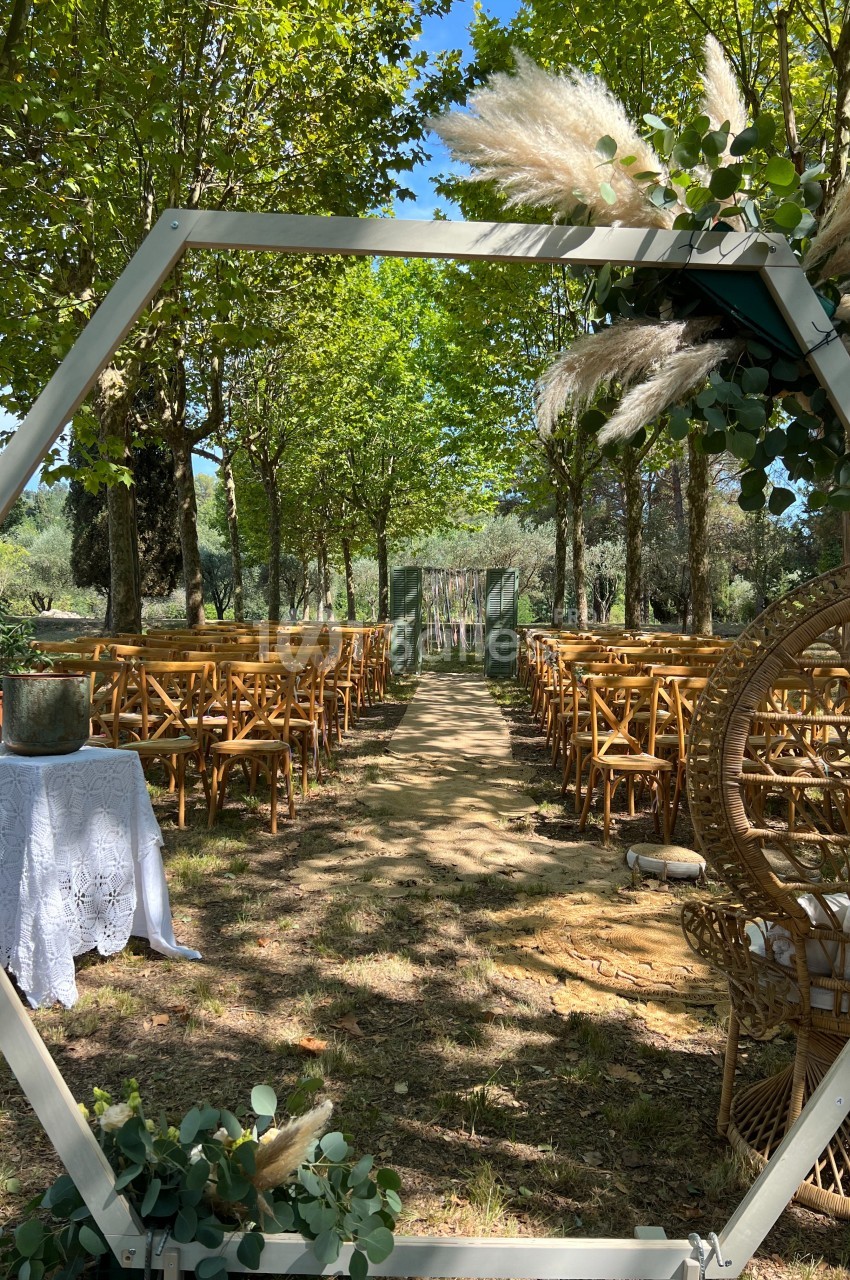 Allée centrale bordée de chaises en bois, décorée de feuillages et de fleurs, menant à une arche dans un cadre naturel.