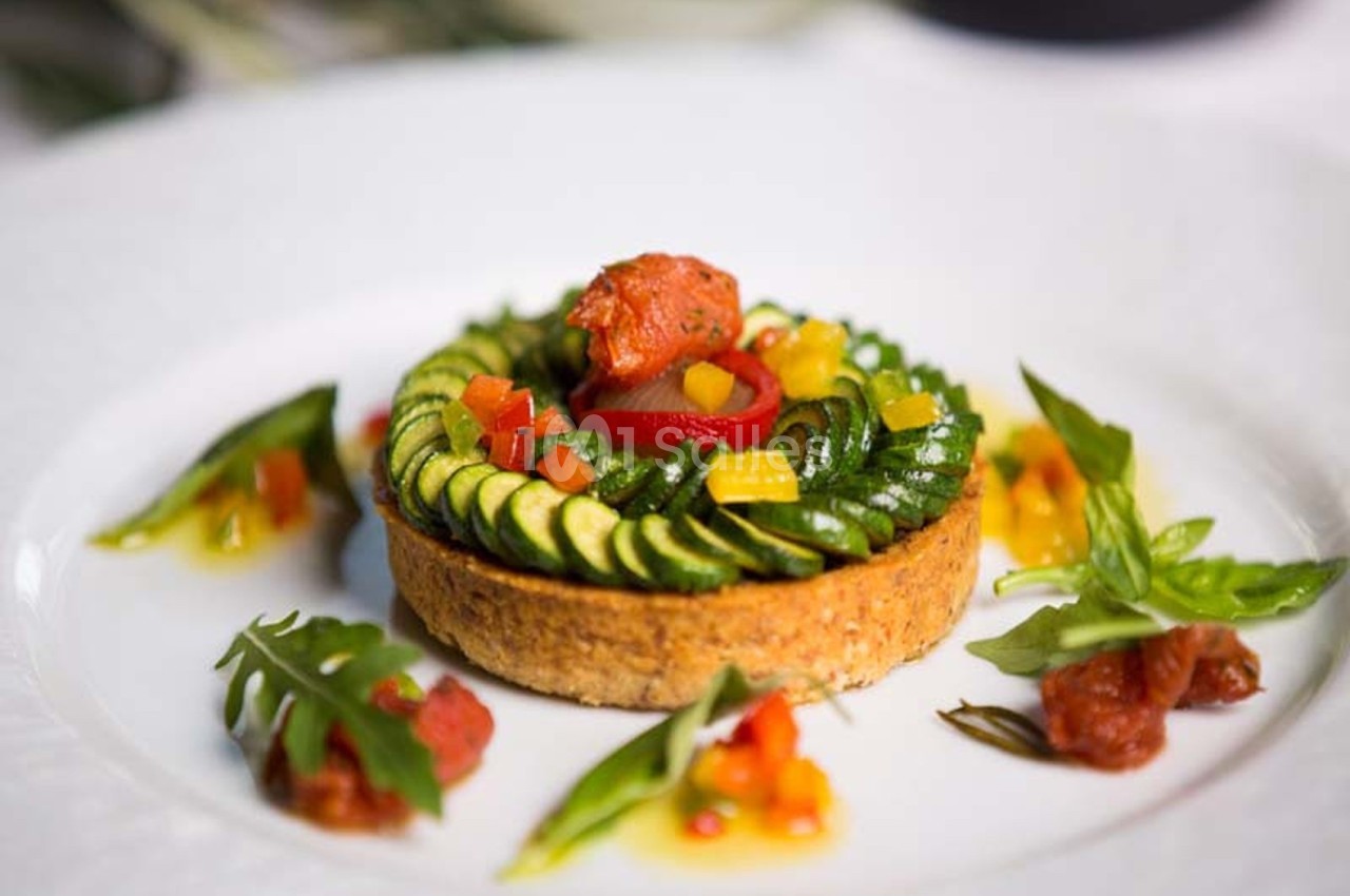 Tartelette salée garnie de courgettes en spirale, légumes colorés et sauce, servie sur une assiette blanche.