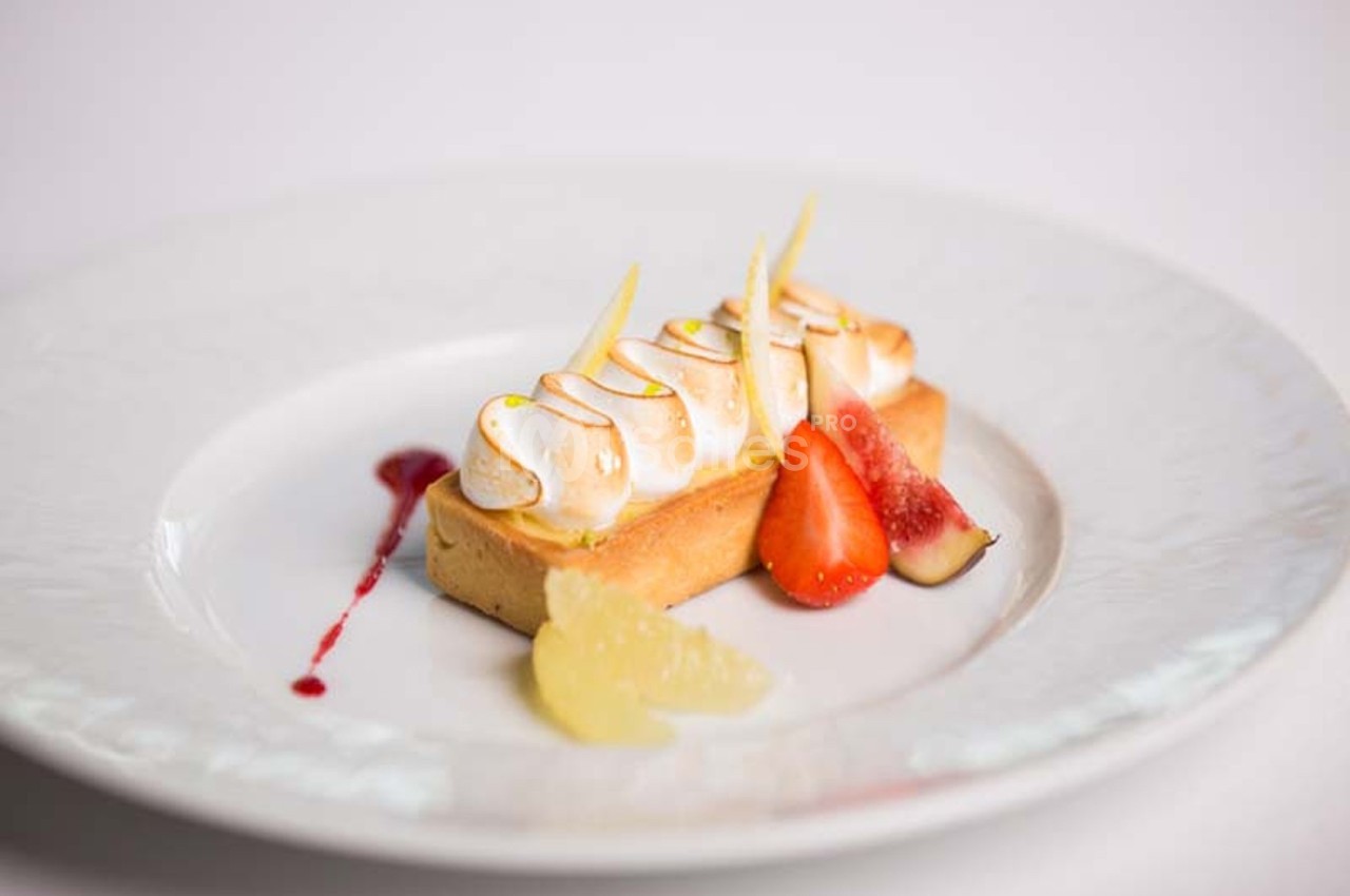 Tarte citron meringuée garnie de fruits frais, servie sur une assiette blanche avec un filet de coulis rouge.