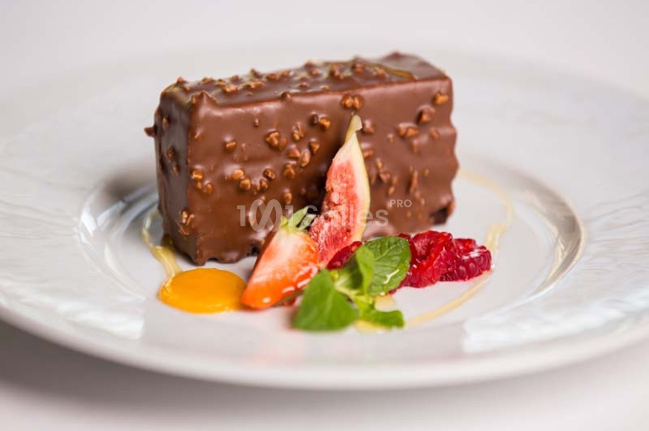 Dessert rectangulaire enrobé de chocolat et noisettes, accompagné de fruits frais et d'une sauce sur une assiette blanche.