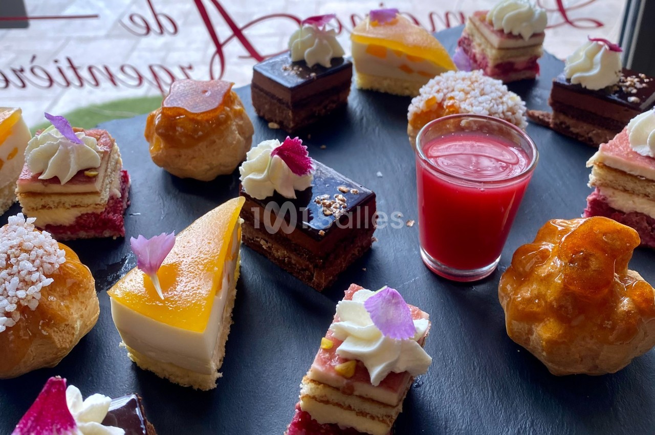 Assortiment de pâtisseries miniatures sur un plateau noir, avec une verrine de coulis rouge au centre.