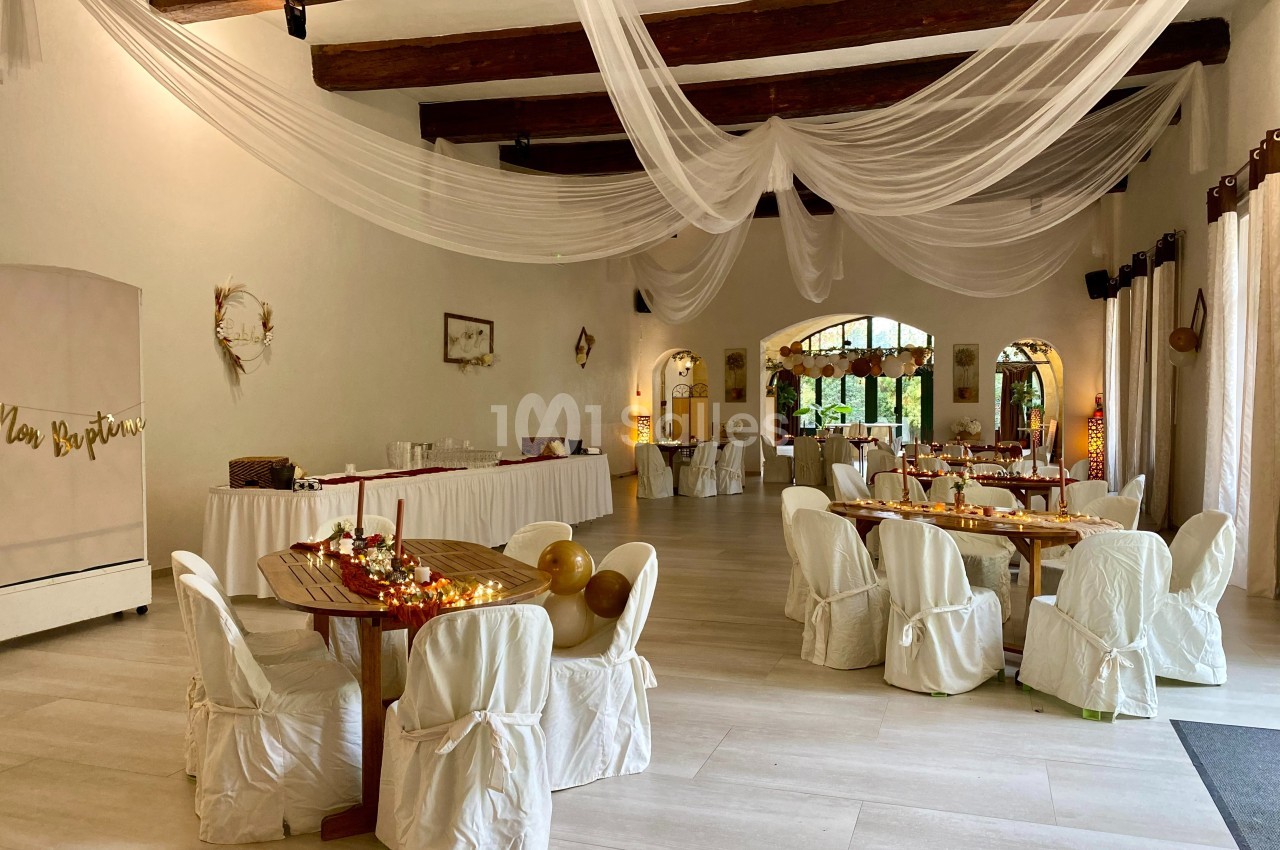 Salle de réception décorée avec des tables rondes, nappes blanches, guirlandes lumineuses et voilages suspendus.