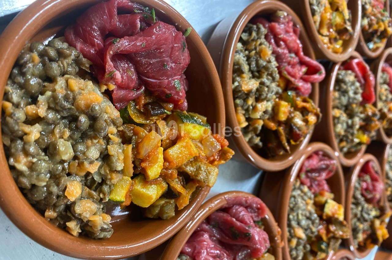 Assiettes en terre cuite contenant des lentilles, légumes sautés et morceaux de viande crue, disposées sur une table.