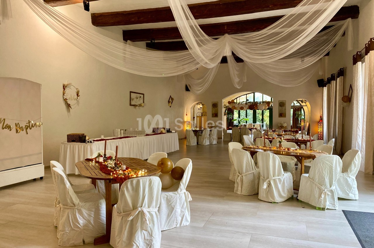 Salle de réception décorée avec des tables rondes, nappes blanches, guirlandes lumineuses et voilages suspendus.