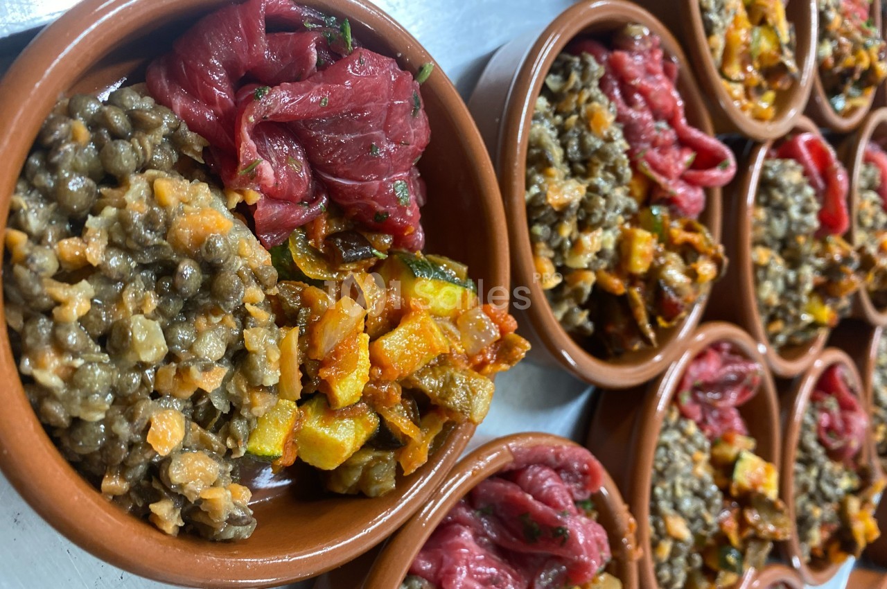 Assiettes en terre cuite contenant des lentilles, légumes sautés et morceaux de viande crue, disposées sur une table.