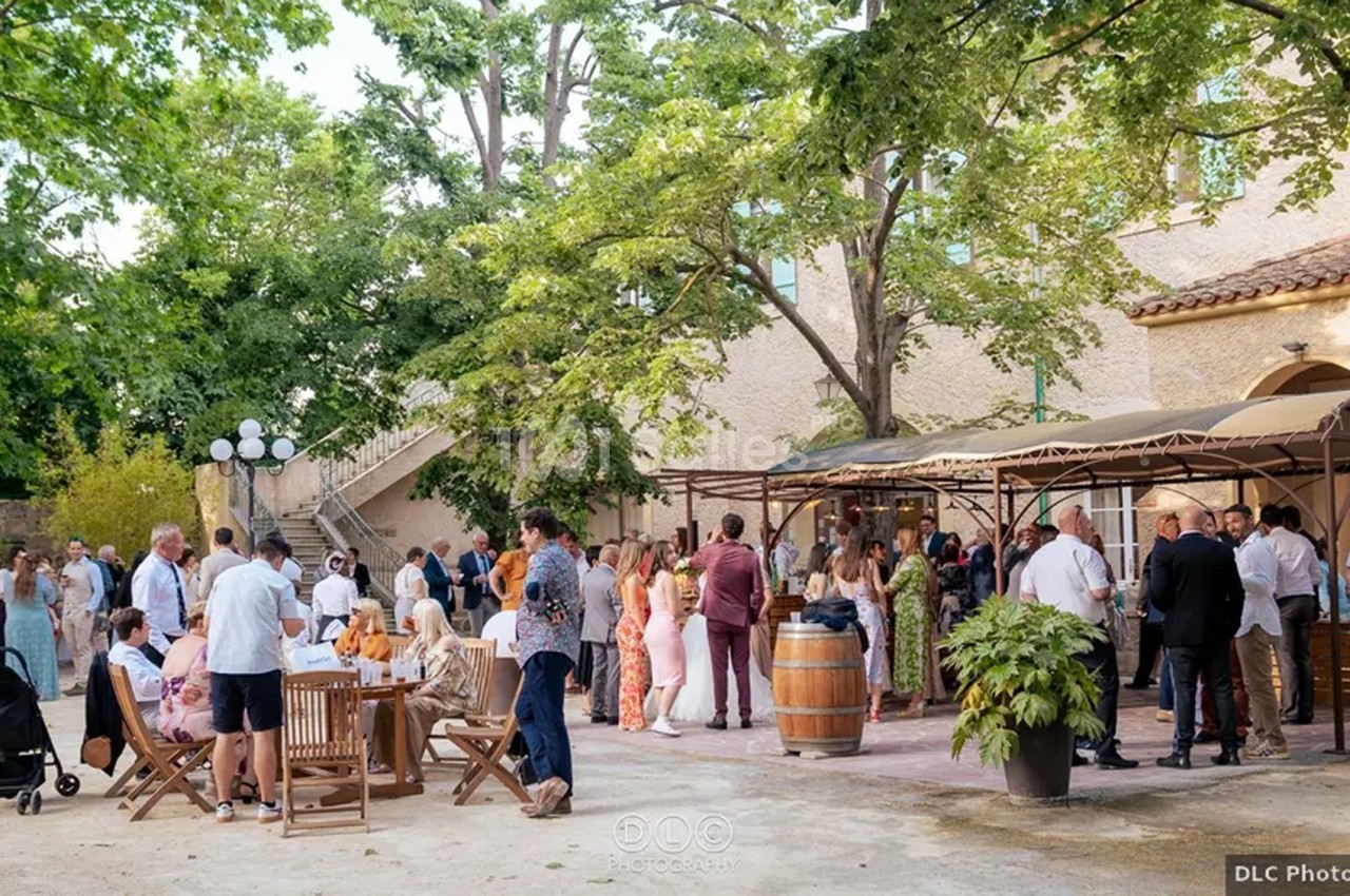 Groupe de personnes rassemblées dans une cour extérieure ombragée, près de tables et d'un bar, lors d'un événement.