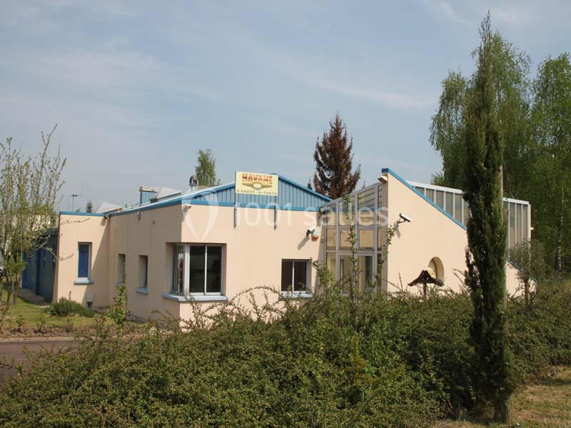 Bâtiment beige avec toit bleu, entouré de végétation et d'arbres, sous un ciel dégagé.