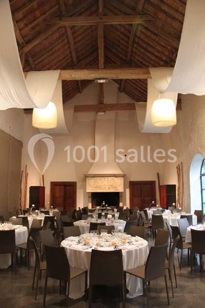 Salle de réception avec tables rondes dressées, chaises modernes, cheminée sculptée et plafond en poutres apparentes.