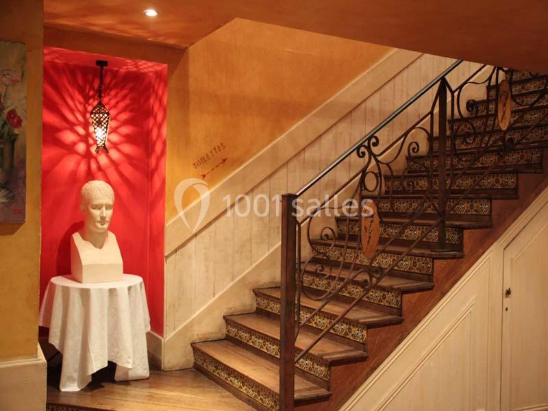 Escalier en bois avec rampe en fer forgé, mur rouge orné d'une applique lumineuse et d'un buste sur une table.
