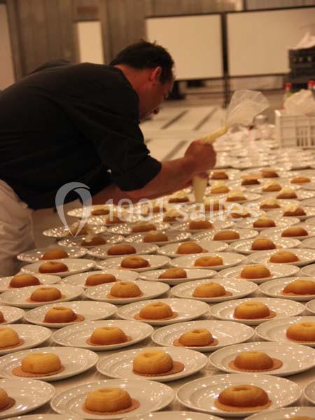 Un chef pâtissier décore des dizaines de desserts alignés sur des assiettes blanches dans une grande cuisine.