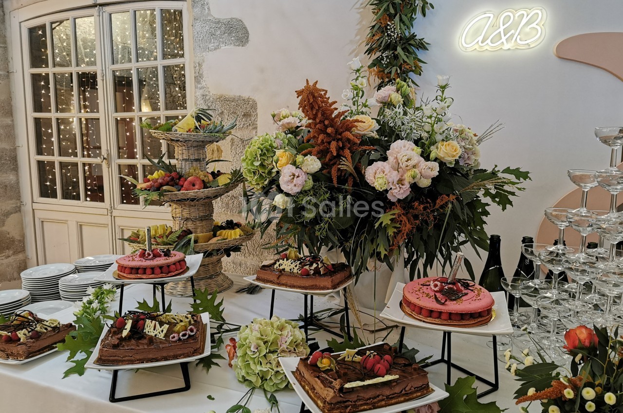 Table décorée avec des fleurs, des gâteaux variés et des fruits, entourée de verres et d'une lumière néon ’A&B’.