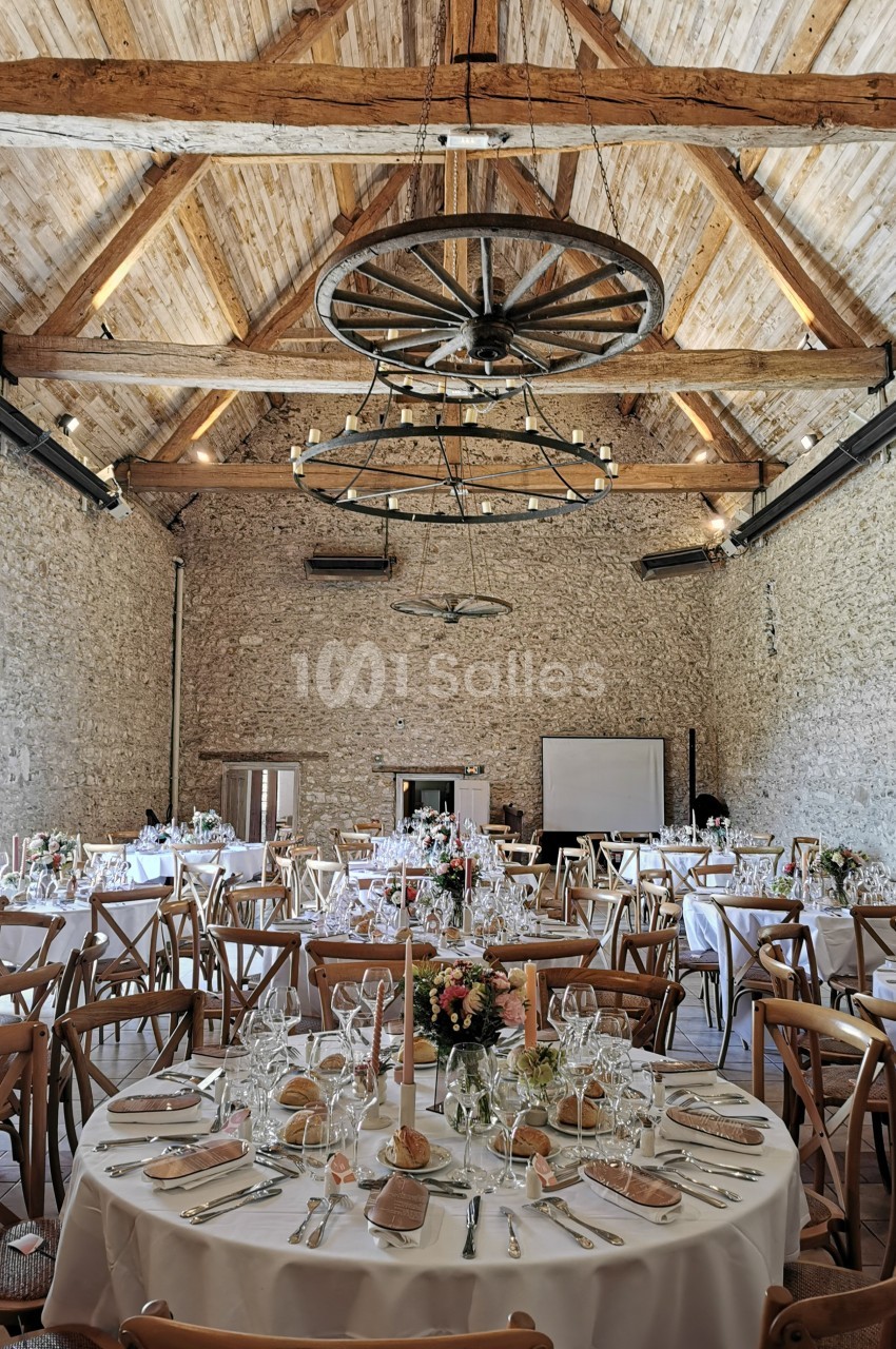 Salle de réception en pierre avec tables rondes dressées, décorations florales et poutres apparentes au plafond.