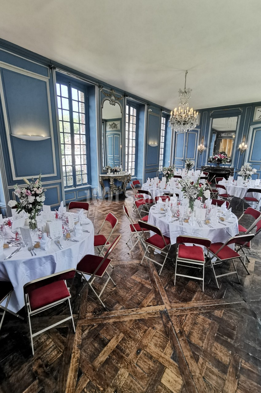 Salle élégante avec murs bleus, tables rondes dressées avec nappes blanches, chaises rouges et décor floral.
