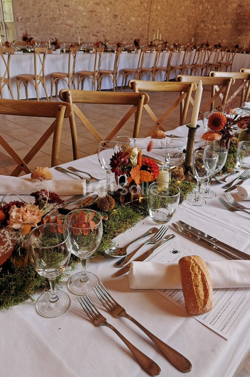 Table décorée pour un repas avec nappes blanches, verres, couverts, bougies et centre de table floral sur mousse.
