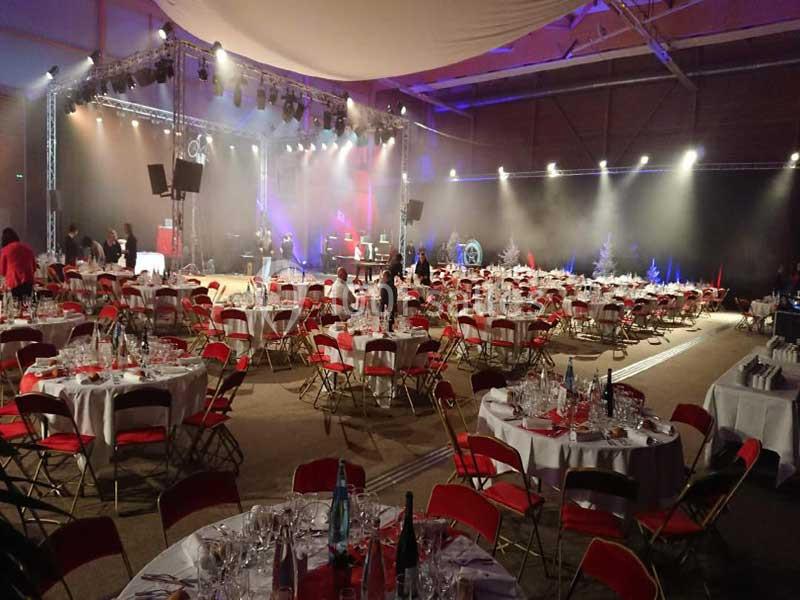 Salle de réception avec tables rondes dressées, chaises rouges et scène éclairée en arrière-plan.