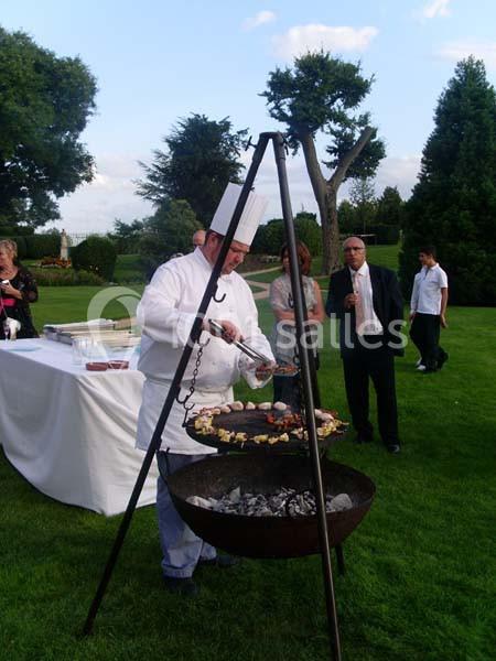 Un chef cuisine des brochettes sur un barbecue suspendu lors d'un événement en plein air dans un jardin.