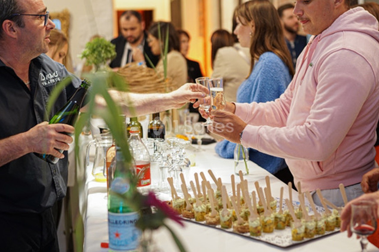 Un serveur remplit un verre de boisson pour un homme lors d'un buffet avec des verrines et des invités en arrière-plan.