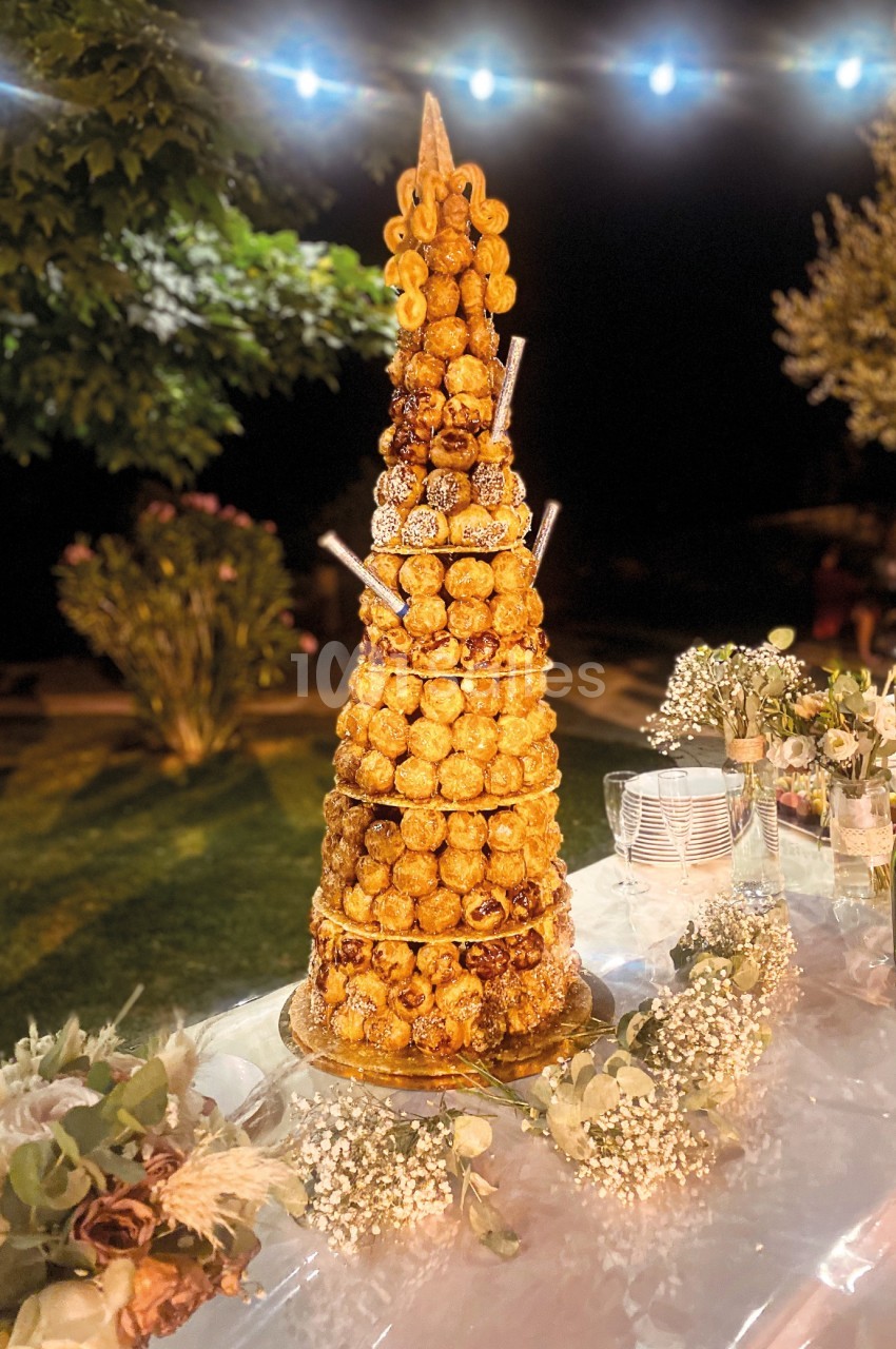 Tour de choux en croquembouche décorée, posée sur une table extérieure éclairée, entourée de fleurs et de vaisselle.