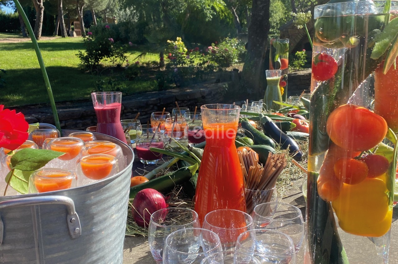 Carafes de jus colorés, légumes et fruits frais disposés sur une table en extérieur, sous un soleil éclatant.