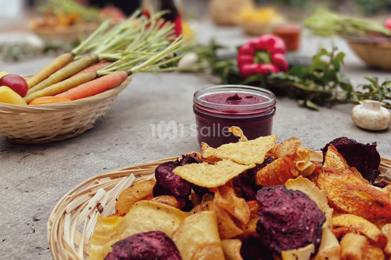 Chips de légumes colorées dans un panier avec un pot de sauce, entourés de légumes frais sur une table en extérieur.