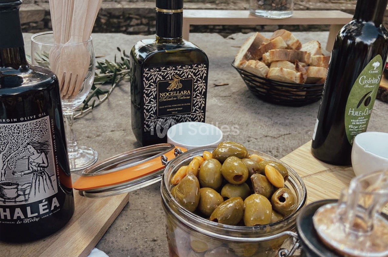 Bocaux d'olives, bouteilles d'huile d'olive et pain disposés sur une table en extérieur.